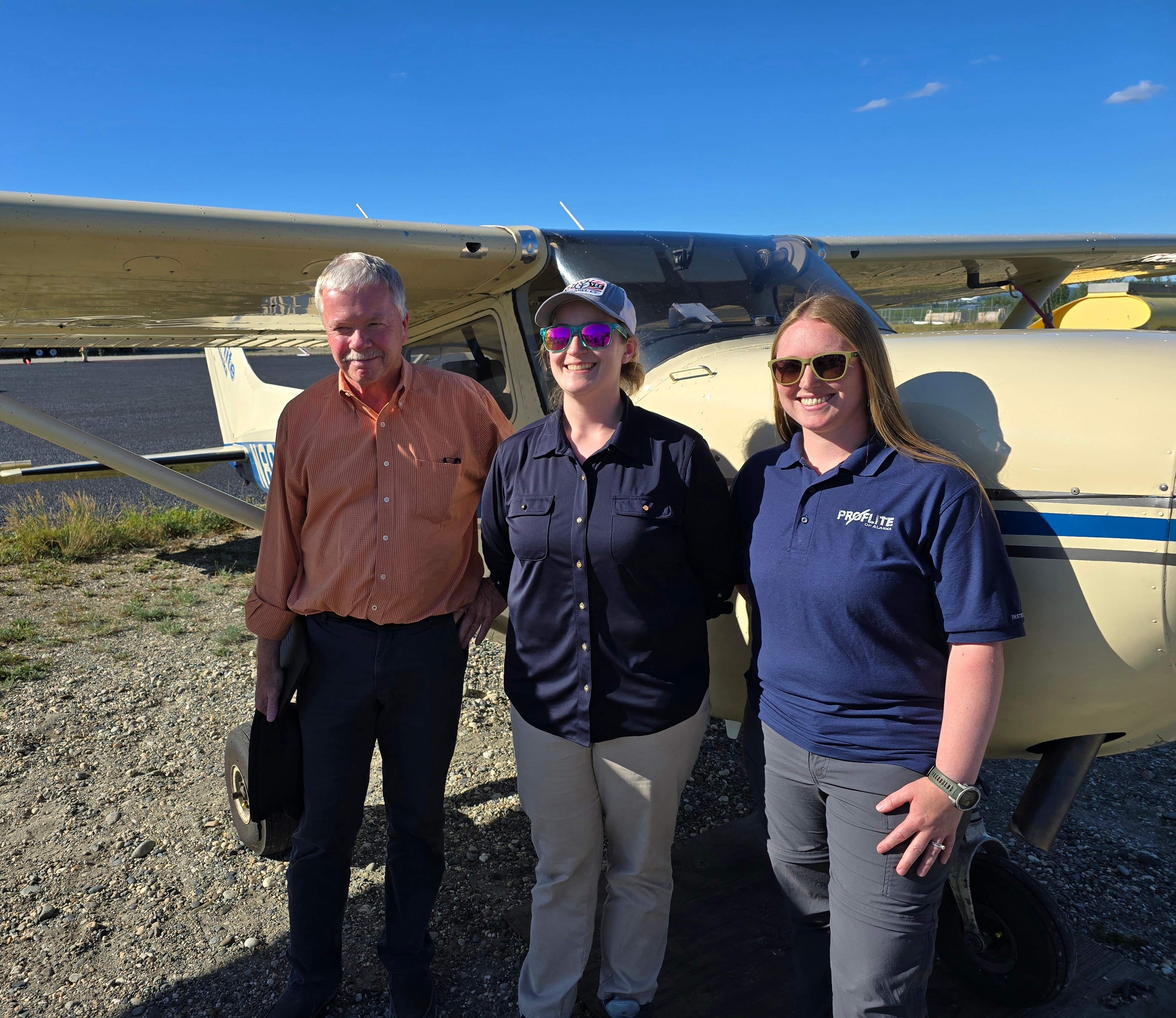 Three people posing in front of a ProFlite of Alaska Cessna on a sunny summer day